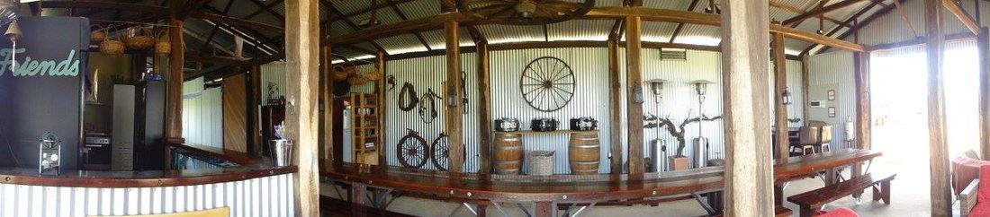 Panoramic view along the inside of the shed showing the bar area, hanging wagon wheel, wine barrels and timber posts down the length of the building.