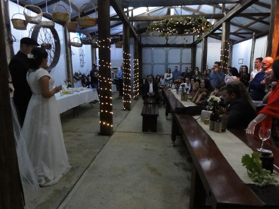 A bride and groom exchange vows in the shed, guests seated along the long jarrah table under fairy lights.